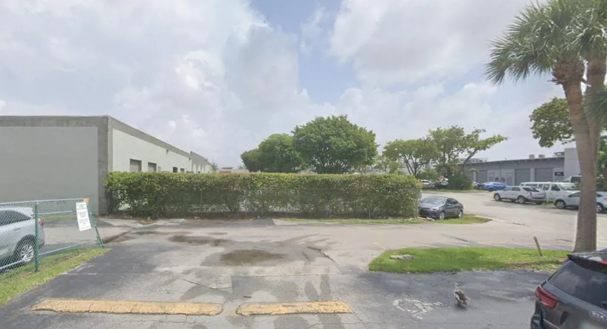 The entrance to the warehouse park on SW 47th Street where Casa Bake operates, with a concrete warehouse, a chain-link fence, and parked cars