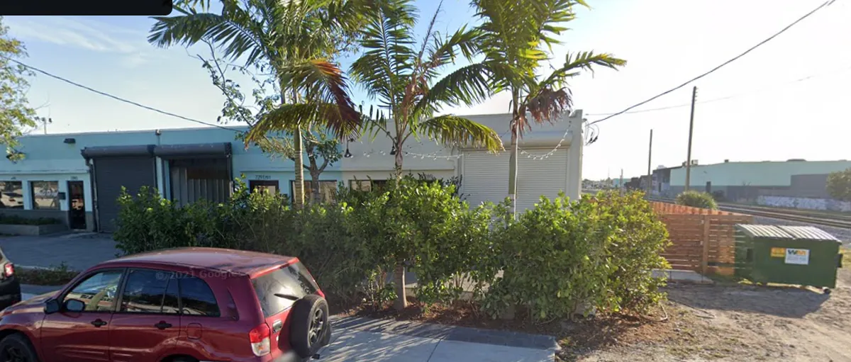 La Natural's low white building behind hedges on NW 2nd Avenue, with the railroad tracks and a dumpster visible to the right